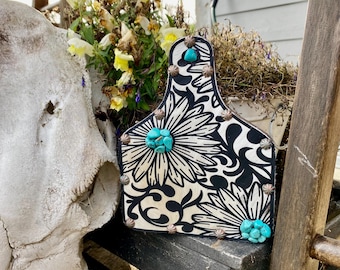 May include: Black and white leather tag with a floral design and turquoise accents. The tag is decorated with silver studs and has a unique shape.