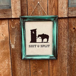May include: A framed sign with a teal and black distressed finish. The sign features a black silhouette of a horse tied to a hitching post, with the words "SHIT & SPLIT" in bold, black letters. The sign is hanging on a wooden wall.