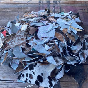 May include: A large pile of assorted leather scraps featuring various textures and patterns, including brown and white cowhide, black and white spotted hide, and solid grey suede pieces with visible cut edges and irregular shapes.
