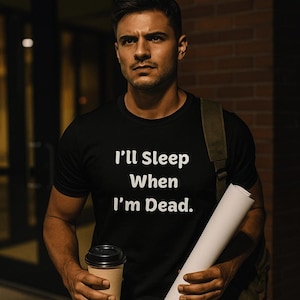 May include: A black short-sleeved t-shirt featuring the white text "I'll Sleep When I'm Dead." centered on the chest. The shirt is worn with a dark olive green backpack, while a paper scroll and a disposable coffee cup are held in the hands.