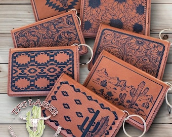 Könnte beinhalten: Eine Sammlung von acht braunen Leder-Schlüsselanhängern mit verschiedenen Designs, darunter ein Kaktus, eine Bergkette, ein Blumenmuster, ein geometrisches Muster und ein Sonnenblumenmuster. Die Schlüsselanhänger sind alle mit schwarzer Tinte graviert und haben einen silbernen Schlüsselring. Die Schlüsselanhänger sind auf einer Holzoberfläche angeordnet. Der Text "TFC LASER WESTERN LASER ENGRAVING" ist auf einem der Schlüsselanhänger sichtbar.