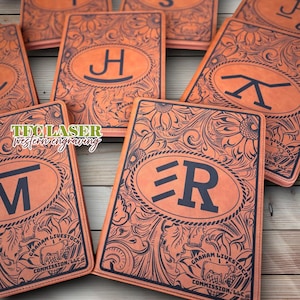 May include: A collection of personalized leather notebooks with intricate floral and initial designs. Each notebook features a unique letter within an oval frame, surrounded by detailed patterns. The notebooks are a warm orange color with dark blue engraved details.