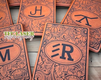 May include: Several orange leather notebooks with dark blue engraved initials and floral patterns. Each notebook has a different initial within an oval. The notebooks are decorated with detailed floral designs and the text "GRAHAM LIVESTOCK COMMISSION, LLC".