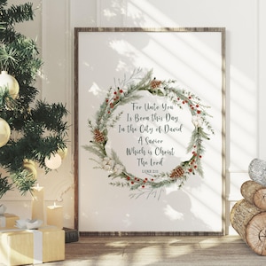 以下が含まれることがあります： クリスマスリースデザインと「For Unto You Is Born this Day In the City of David A Savior Which is Christ The Lord Luke 2:11」のテキストがプリントされた額入りのプリント。装飾されたクリスマスツリーと包装されたギフトの近くにプリントが表示されています。