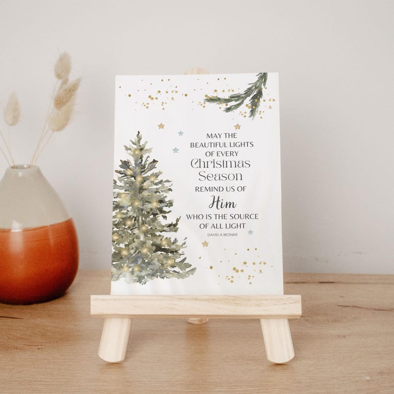 May include: A Christmas card displayed on a miniature wooden easel. The card shows a watercolour illustration of a Christmas tree with lights and the text: "May the beautiful lights of every Christmas season remind us of Him."