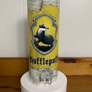 May include: A yellow and gold tumbler with a black badger and the words "Hufflepuff" in a black banner. The background is a vintage parchment with text and a crest.