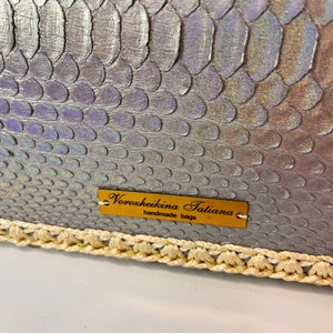 Puede incluir: Un bolso de mano plateado iridiscente con ribete de ganchillo. El bolso tiene una etiqueta dorada que dice "Voroxheikina Tatiana handmade bags".