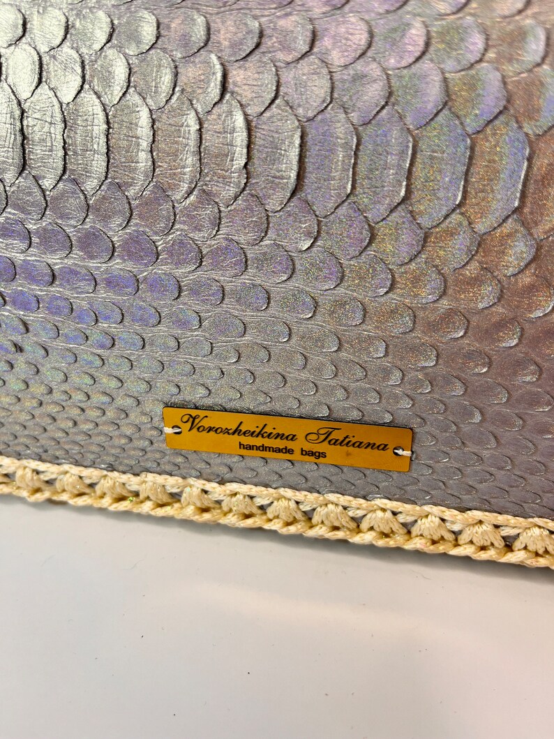 Puede incluir: Un bolso de mano plateado iridiscente con ribete de ganchillo. El bolso tiene una etiqueta dorada que dice "Voroxheikina Tatiana handmade bags".