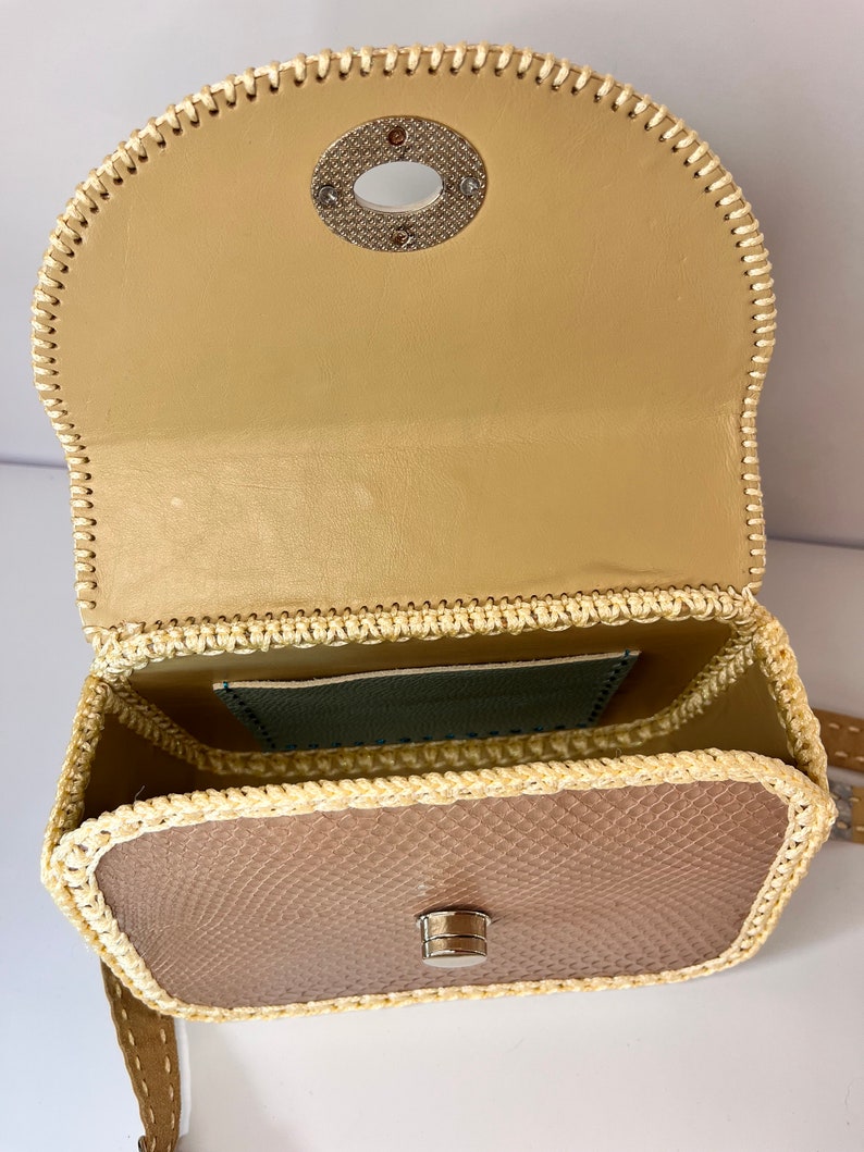 Puede incluir: Un peque&ntilde;o bolso de mano de cuero beige con ribete tejido y cierre de metal. El bolso tiene una solapa superior con un anillo de metal y un peque&ntilde;o bolsillo en el interior.