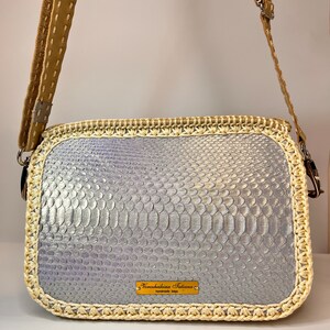 Puede incluir: Un bolso bandolera de piel de serpiente plateada con ribete de crochet crema y una correa de cuero marr&oacute;n. El bolso tiene una etiqueta de metal dorado que dice "Noretshekina Tatiana handmade bags".