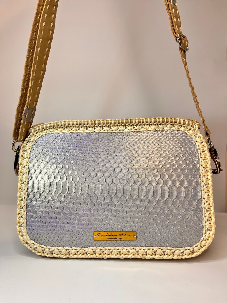 Puede incluir: Un bolso bandolera de piel de serpiente plateada con ribete de crochet crema y una correa de cuero marr&oacute;n. El bolso tiene una etiqueta de metal dorado que dice "Noretshekina Tatiana handmade bags".