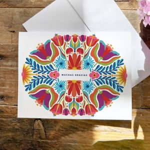 May include: A white card with a colorful floral design in shades of red, orange, yellow, green, blue, and purple. The card has the text "MUCHAS GRACIAS" in the center.