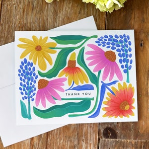 Puede incluir: Una tarjeta blanca con un dise&ntilde;o floral colorido. La tarjeta presenta flores rosas, amarillas, azules y rojas con hojas verdes. El texto "THANK YOU" est&aacute; impreso en negro en la tarjeta.