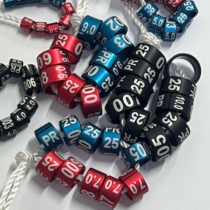 Puede incluir: Surtido de anillos de plástico de colores con números y letras blancas. Los anillos se utilizan para marcar e identificar artículos. Los anillos son de color rojo, azul y negro.