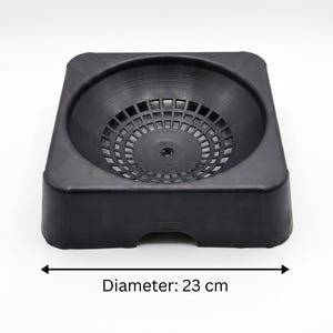 May include: A black plastic bowl-shaped stand with a square base and a circular, grid-patterned interior. The object features a central drainage hole and a small arched cutout at the bottom of one side. The total diameter measures 9.06 inches.