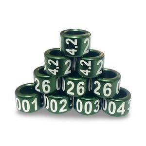 Puede incluir: Una pirámide de anillas verdes de metal para aves con números y marcas blancas. Las anillas están apiladas, mostrando números como "4.2 2" y "26 001". Las anillas son cilíndricas y tienen un brillo metálico.