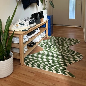 May include: A wooden shoe rack with two shelves, filled with white and black sneakers. A green and white patterned rug sits in front of the rack. Two potted plants are on either side of the rack.