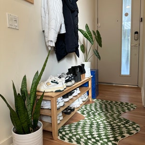 May include: A wooden shoe rack with multiple shelves, filled with white and black trainers. The rack is placed in a hallway with a green patterned rug, a potted snake plant, and a coat rack with jackets hanging on it.