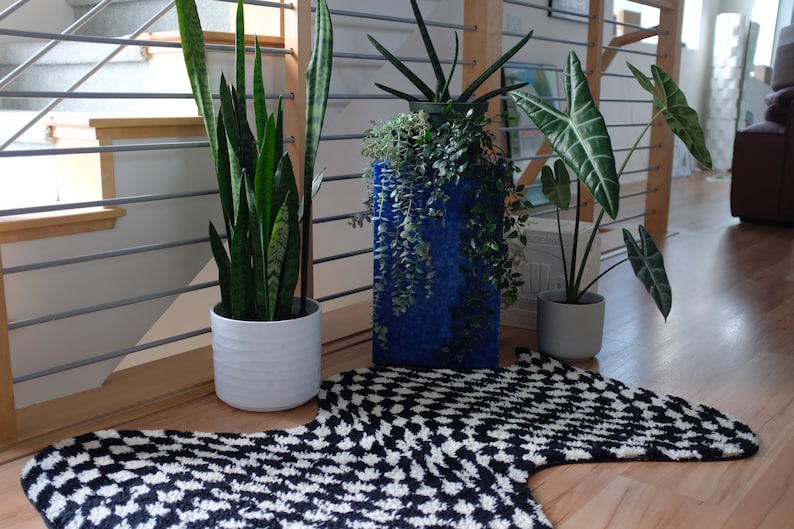 May include: Three potted plants on a black and white patterned rug. The plants are a snake plant in a white pot, a leafy plant in a blue pot, and a large-leafed plant in a grey pot.
