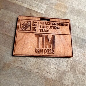 May include: A wooden name badge with the Home Depot logo and the text "MET MERCHANDISING EXECUTION TEAM". The name "TIM" and the code "DEM D332" are also engraved on the badge. The badge is rectangular with a slot at the top.