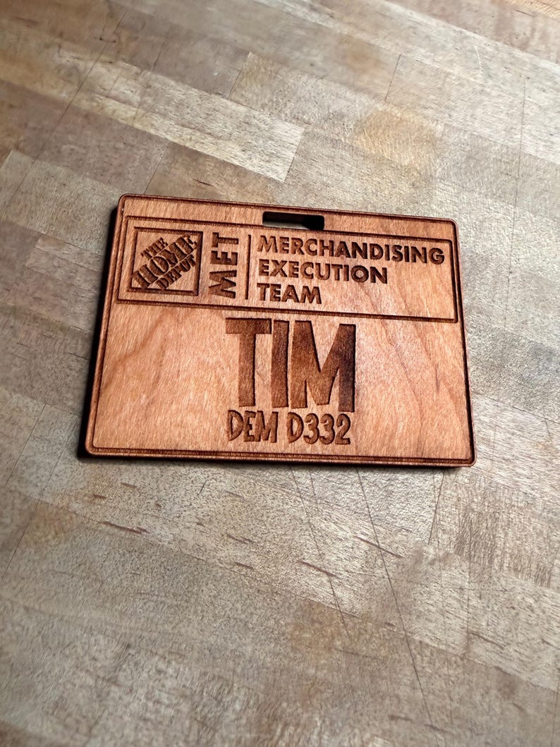 May include: A wooden name badge with the Home Depot logo and the text "MET MERCHANDISING EXECUTION TEAM". The name "TIM" and the code "DEM D332" are also engraved on the badge. The badge is rectangular with a slot at the top.