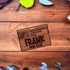 May include: A wooden name tag with the text "FRANK" and "DEM D242" is displayed on a wooden surface, surrounded by tools like wrenches, pliers, and a yellow tape measure. The tag also includes "MERCHANDISING EXECUTION TEAM" and the Home Depot logo.