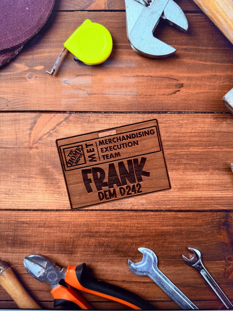 May include: A wooden name tag with the text "FRANK" and "DEM D242" is displayed on a wooden surface, surrounded by tools like wrenches, pliers, and a yellow tape measure. The tag also includes "MERCHANDISING EXECUTION TEAM" and the Home Depot logo.
