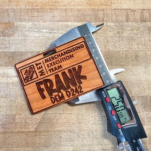 May include: A wooden name tag with the text "FRANK DEM D242" and "MERCHANDISING EXECUTION TEAM" is measured with a digital caliper. The tag also features the Home Depot logo. The caliper reads 2.50 inches.