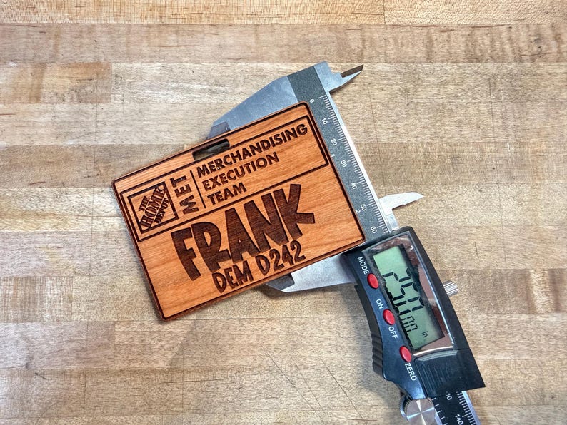 May include: A wooden name tag with the text "FRANK DEM D242" and "MERCHANDISING EXECUTION TEAM" is measured with a digital caliper. The tag also features the Home Depot logo. The caliper reads 2.50 inches.