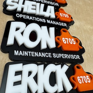 May include: A collection of black and white name badges with the names Marilyn, Sheila, Ron, and Erick. Each badge has a white name and title, with an orange apron-shaped accent and the number 6705.