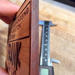 May include: A rectangular wooden sign with the name "FRANK" and other text, including "MERCHANDISING EXECUTION TEAM" and a phone number. The sign is measured with a digital caliper, showing a thickness of approximately 0.35 inches.