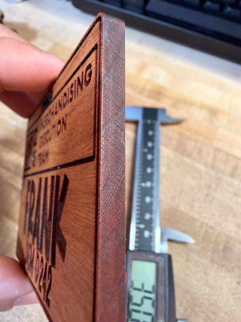 May include: A rectangular wooden sign with the name "FRANK" and other text, including "MERCHANDISING EXECUTION TEAM" and a phone number. The sign is measured with a digital caliper, showing a thickness of approximately 0.35 inches.