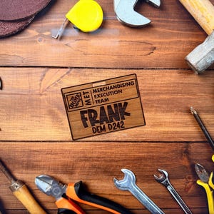 May include: A collection of tools, including wrenches, a hammer, and pliers, arranged on a wooden surface. A wooden name tag with the name "FRANK" and the text "MERCHANDISING EXECUTION TEAM" is also present. The tools are in various colors.