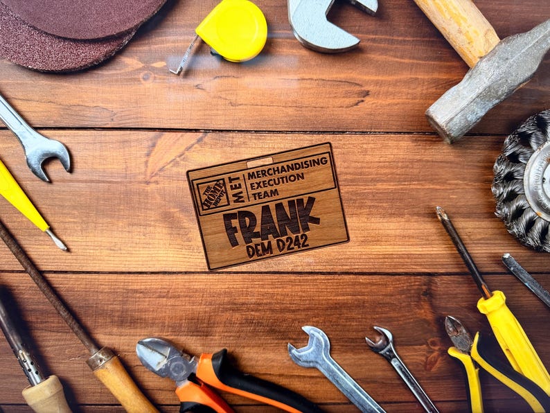 May include: A collection of tools, including wrenches, a hammer, and pliers, arranged on a wooden surface. A wooden name tag with the name "FRANK" and the text "MERCHANDISING EXECUTION TEAM" is also present. The tools are in various colors.