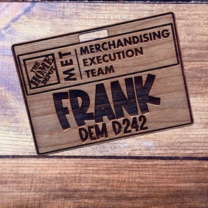 May include: A wooden name tag with the Home Depot logo, the text "MET MERCHANDISING EXECUTION TEAM", and the name "FRANK" with "DEM D242" below it. The tag is rectangular with a hole at the top and is set against a wooden background.