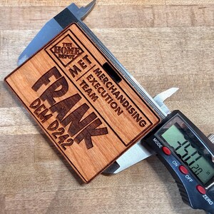 May include: A wooden name tag with the name "FRANK" engraved in large letters. The tag also includes the text "THE HOME DEPOT", "MET MERCHANDISING EXECUTION TEAM", and "DEM D242". The tag is being measured with a digital caliper, showing a measurement of 3.50 inches.