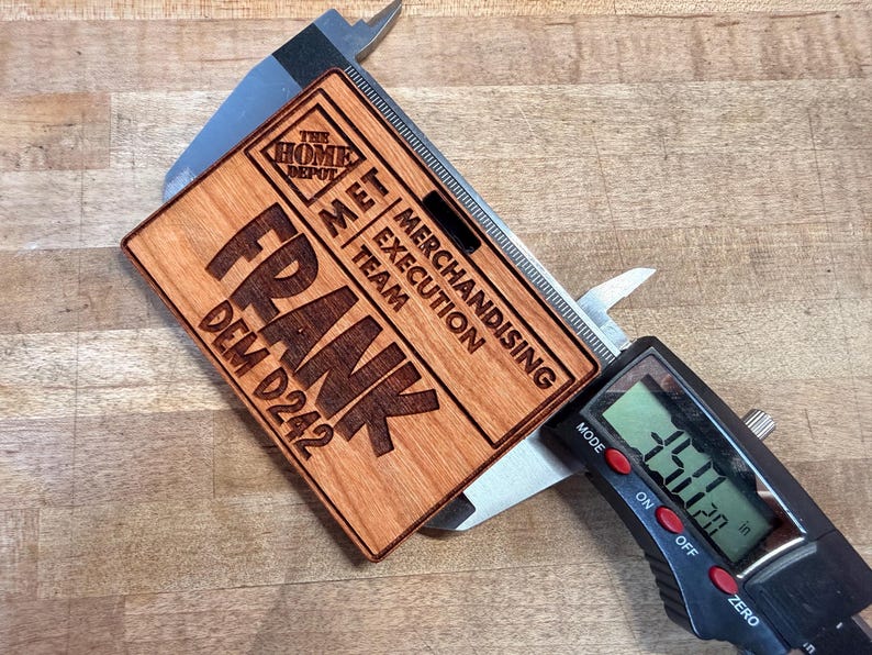 May include: A wooden name tag with the name "FRANK" engraved in large letters. The tag also includes the text "THE HOME DEPOT", "MET MERCHANDISING EXECUTION TEAM", and "DEM D242". The tag is being measured with a digital caliper, showing a measurement of 3.50 inches.