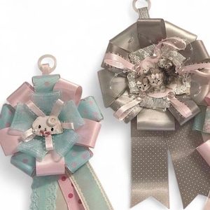 May include: Two decorative ribbon rosettes. The left rosette features light blue and pink polka-dot ribbons with a central white cat-shaped charm. The right rosette is made of metallic taupe ribbons with a central image of three kittens.
