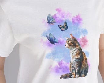 May include: White t-shirt featuring a watercolor design of a cat and butterflies. The cat is brown and orange, with blue and purple butterflies floating around it. The shirt is a classic crew neck style.