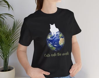 以下が含まれることがあります： 青い地球儀の上に白いシルエットの猫が座っている黒いTシャツ。地球儀の下に緑色の文字で「Cats rule the world」と書いてある。