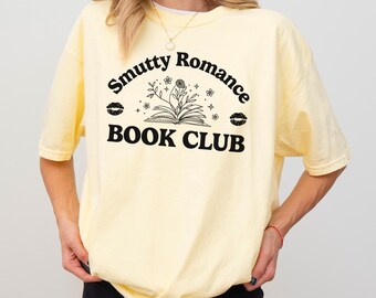 Op de afbeelding: Een geel T-shirt met de tekst "Smutty Romance Book Club" in zwart. De tekst staat in een boog met een gestileerd open boek en bloemen in het midden.
