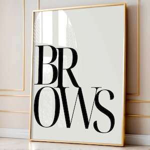 May include: A framed art print featuring the word "BROWS" in large, bold, black lettering on a light beige backdrop. The frame is a thin, gold colour. The artwork is displayed on a wall with gold trim.