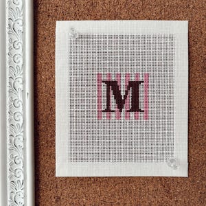 May include: A square piece of white needlepoint canvas featuring a centered dark brown letter M. The letter is layered over a series of vertical light pink stripes. The canvas is secured with two clear push pins to a brown cork board surface.