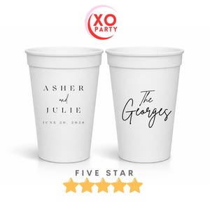 May include: Two white party cups with black text. One cup reads "Asher and Julie" with the date "June 20, 2028". The other cup reads "The Georges". The cups are for a wedding or party.