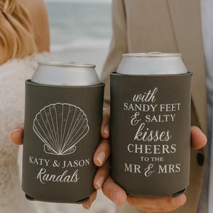 May include: Two grey can coolers with white text and designs. One has a seashell graphic and the names "Katy & Jason Randals." The other says "With sandy feet & salty kisses, cheers to the Mr & Mrs."