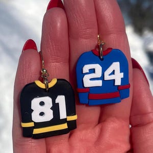 May include: Hockey jersey earrings in black and blue. The black jersey has the number 81 in white, and the blue jersey has the number 24 in white. Both earrings have gold-colored hooks and are designed to resemble miniature hockey jerseys.