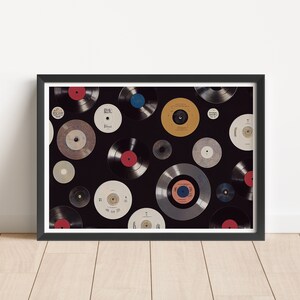 May include: A framed print showing a selection of vinyl records against a black backdrop. The records have various label colours, including red, blue, and yellow. The print is in a black frame, displayed against a white wall and wooden floor.