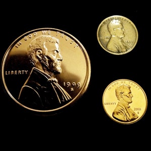 Puede incluir: Tres monedas de un centavo de dólar estadounidense de color dorado con el perfil de Abraham Lincoln en el anverso. Las monedas están fechadas en 1999, 2001 y 1919.