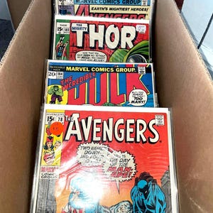 May include: A cardboard box filled with vintage Marvel Comics. The comics feature titles like "Thor," "The Incredible Hulk," and "The Avengers." The covers display vibrant colors and classic comic book art.