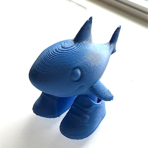May include: A 3D printed blue shark figurine with a visible layered texture. The shark has a rounded body, a dorsal fin, a tail fin, and two side fins. It is standing on two large, rounded blue feet that are also 3D printed with visible layers.